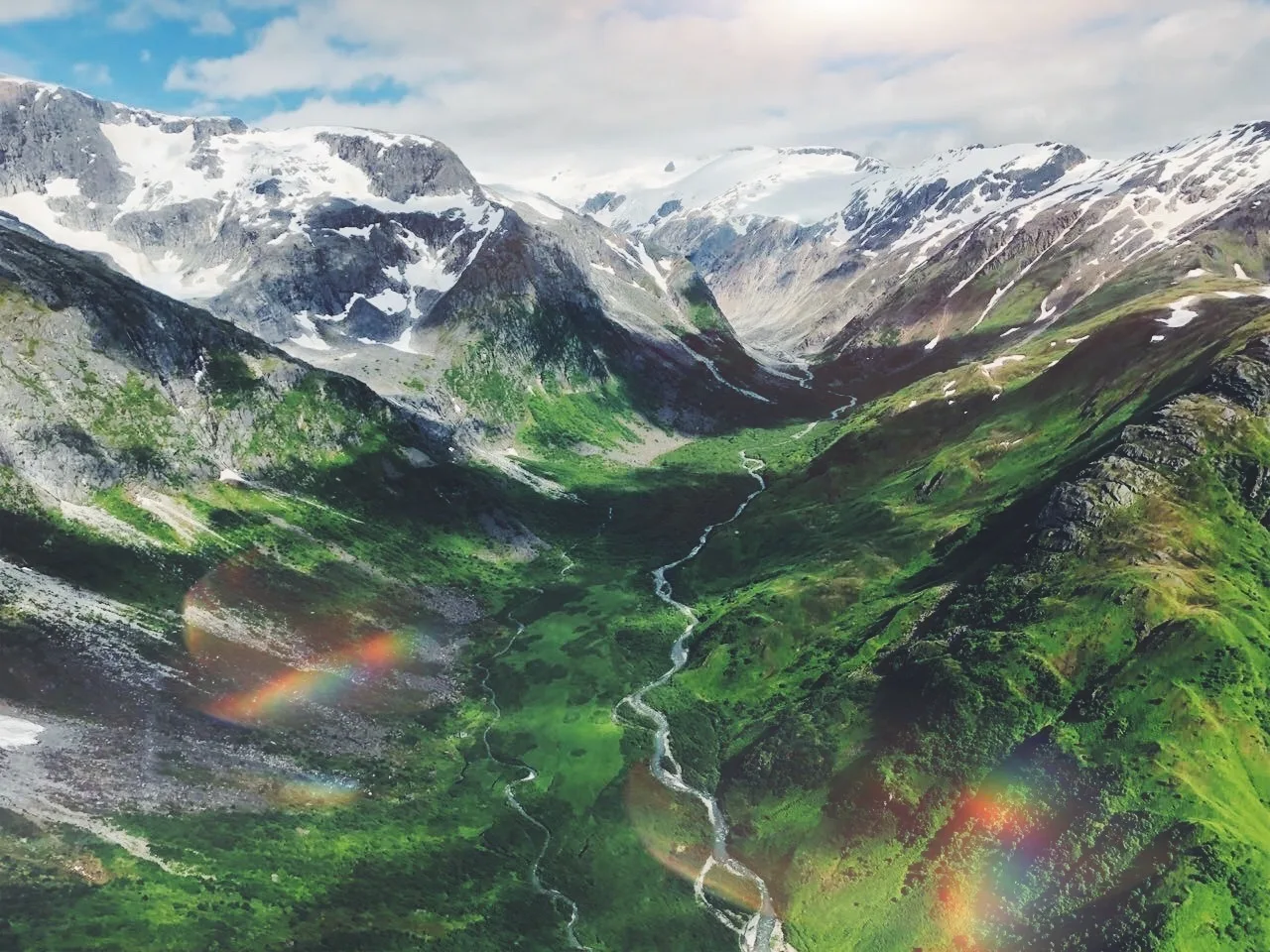 Aerial view of a lush green valley surrounded by snow-capped mountains, with a winding river flowing through the landscape, under a partly cloudy sky.