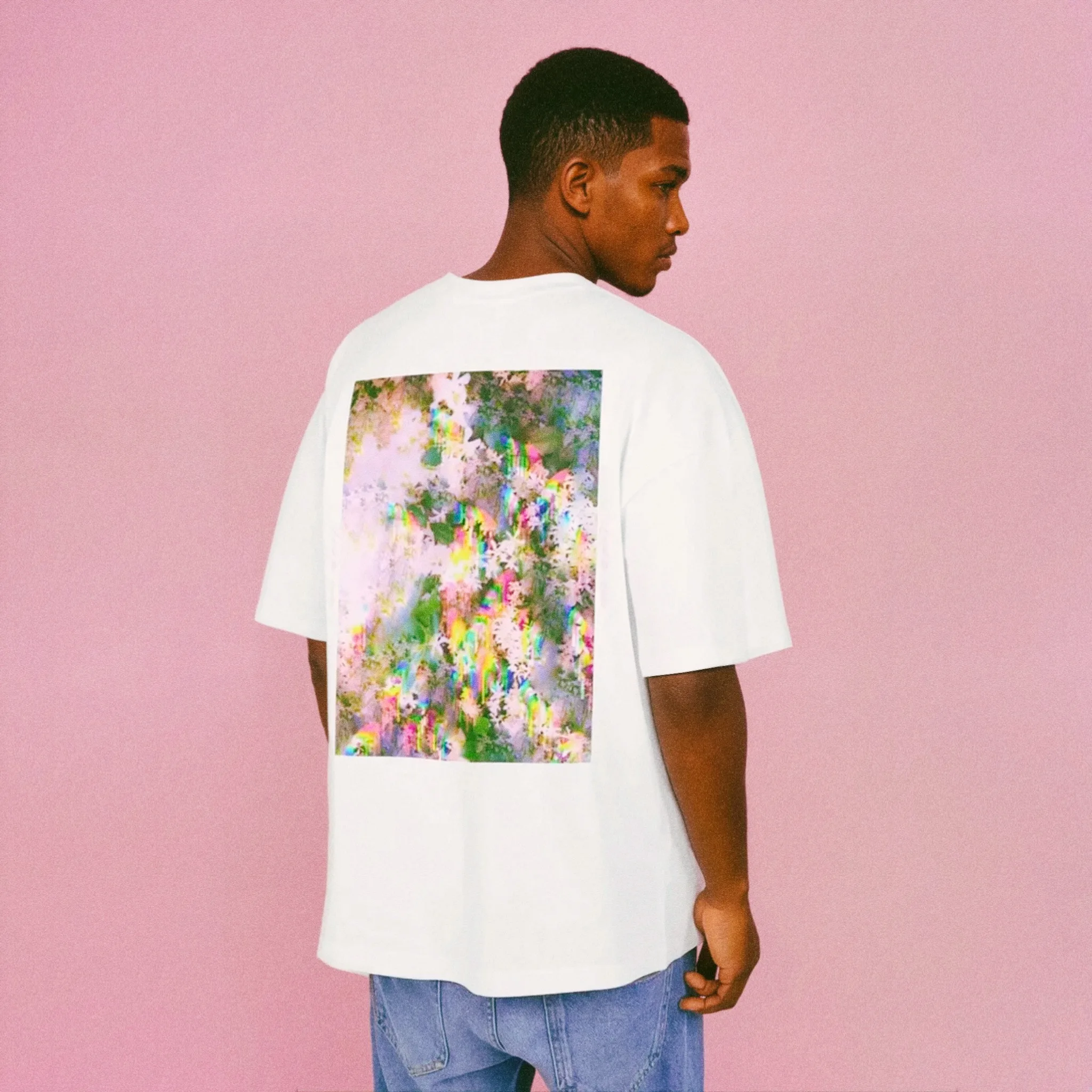 A man wearing a white oversized T-shirt with a colorful abstract print on the back, standing against a pink background.