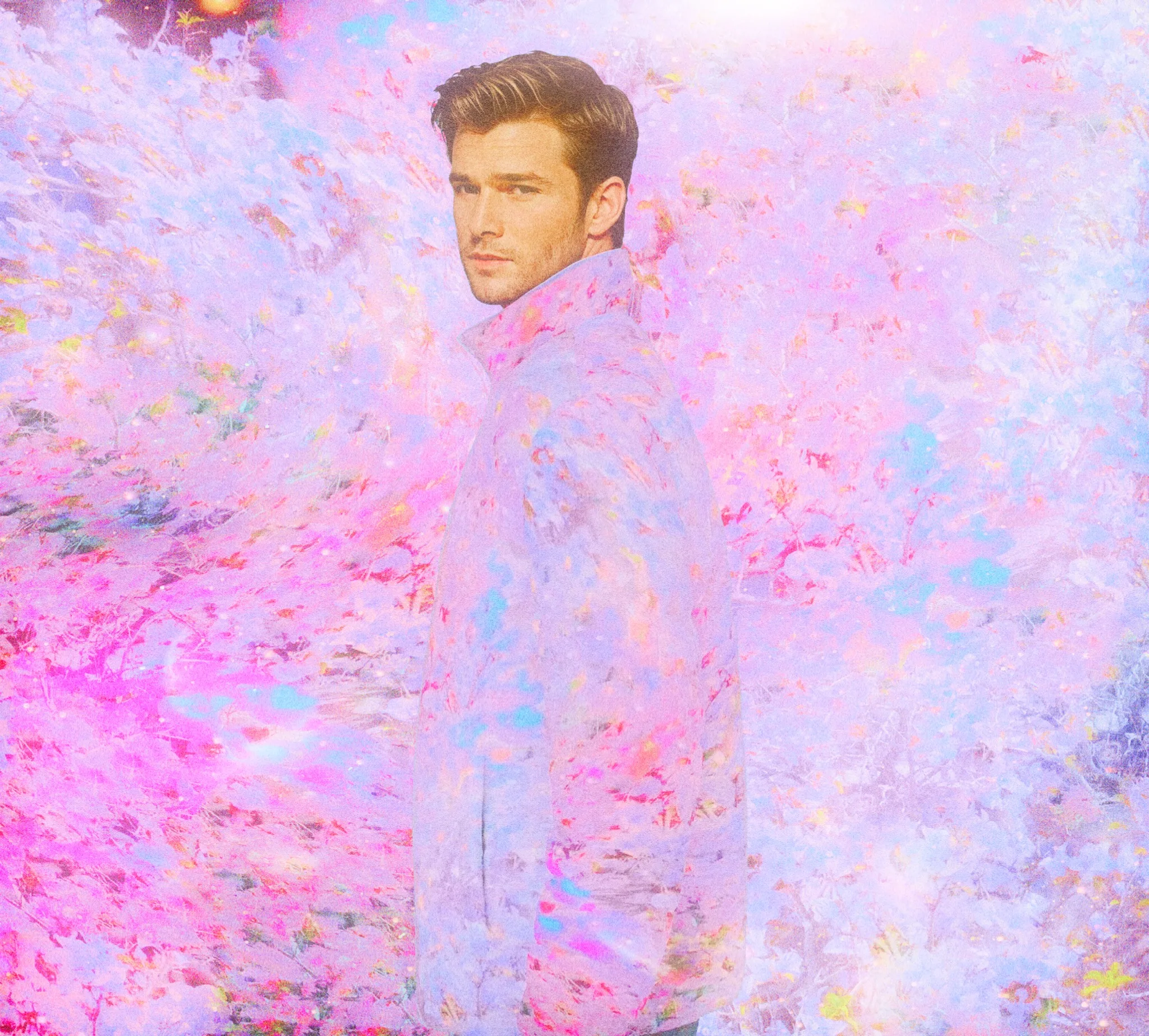 A man wearing a pastel jacket stands against a vibrant, colorful background filled with soft pink and blue tones, suggesting a dreamlike scenery.