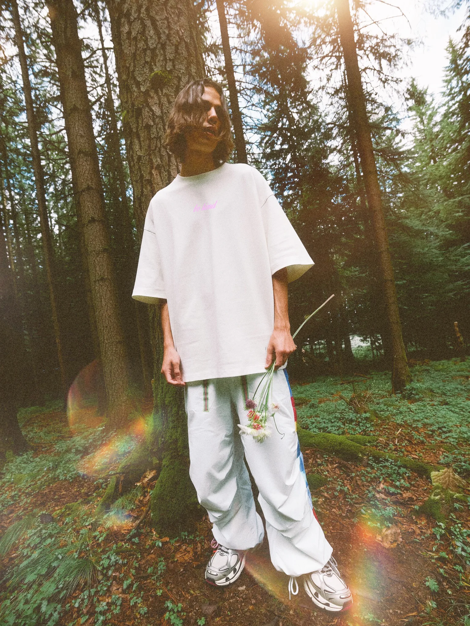 A person stands in a forest, wearing a white oversized t-shirt and light-colored baggy pants. They hold a small bouquet of wildflowers in one hand.