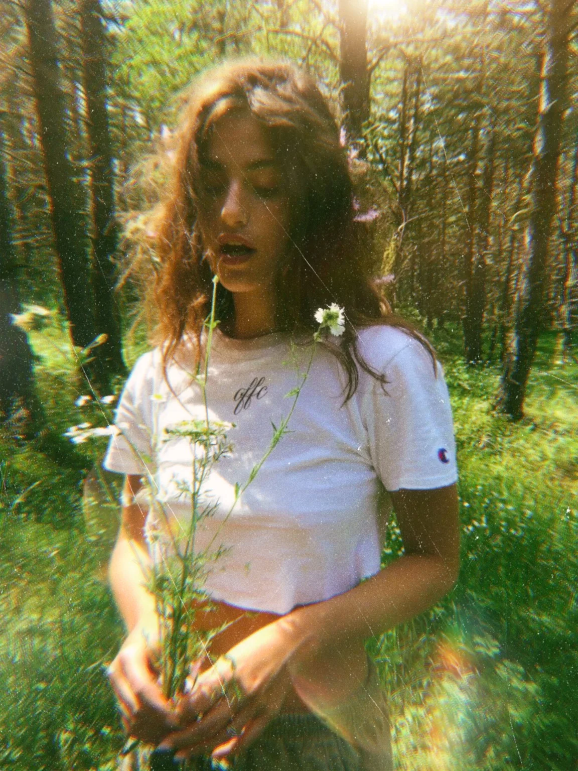 A young woman stands in a forest holding a flower. She has wavy hair and wears a white cropped T-shirt with a small logo. Sunlight filters through the trees, creating a soft, dreamlike atmosphere.