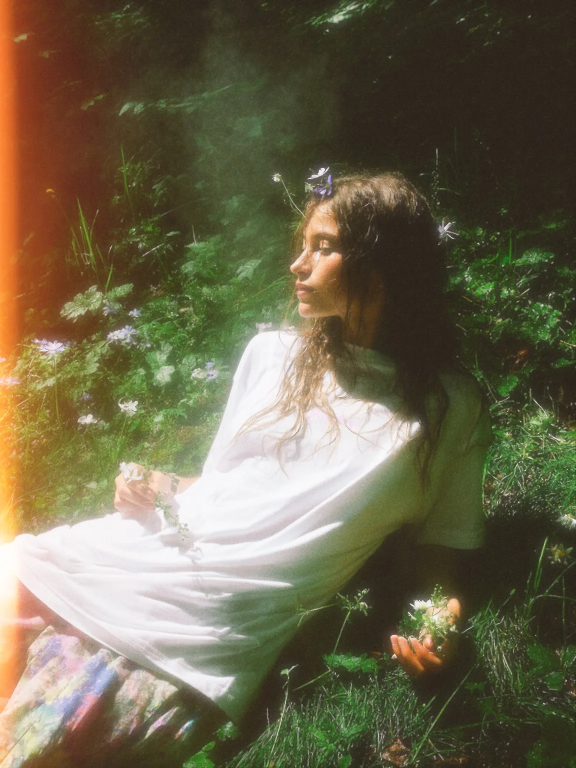 A model with long hair adorned with flowers, wearing a white tee, relaxed on a grassy ground filled with wildflowers in a forest setting.