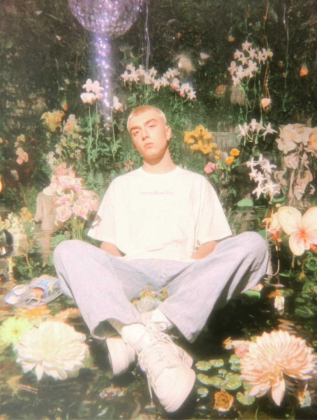 A person with a short haircut seated on a colorful floral background, wearing a white t-shirt and light blue jeans.