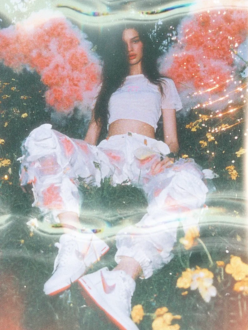 A model sitting amidst flowers, wearing a white cropped top and floral baggy pants, with red and white sneakers. Soft lighting and a peach smoke effect create a dreamy atmosphere.