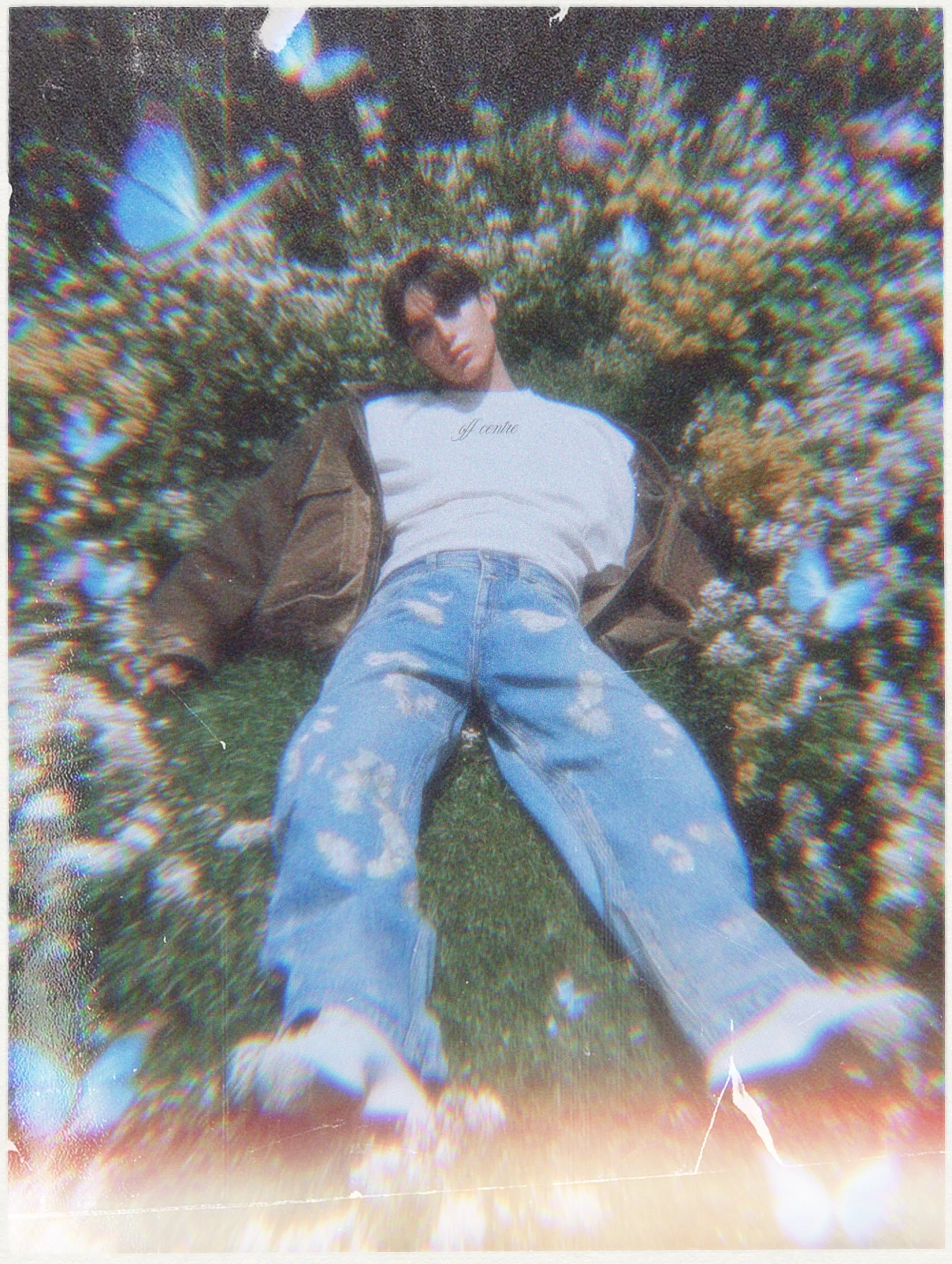 A person lounging on grass surrounded by flowers, wearing a white sweatshirt with the text 'off centre' and baggy jeans. The image has a dreamy, slightly distorted effect with blue butterflies around.