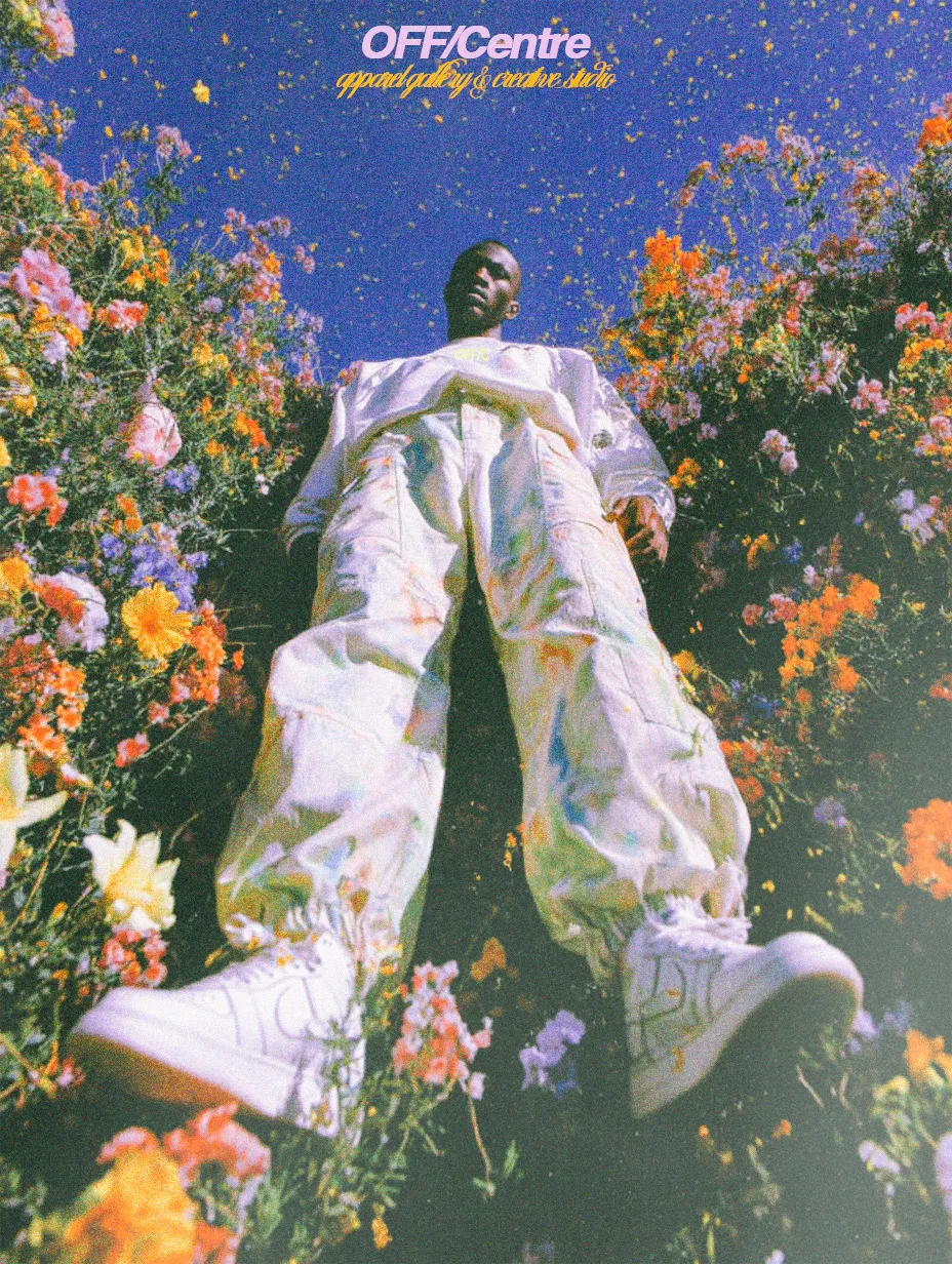 A person standing among colorful flowers, wearing white apparel with a relaxed fit, looking upward under a bright sky.