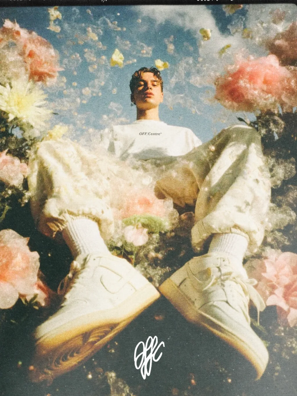 A person sitting among colorful flowers, wearing a white OFF/Centre t-shirt and styled with light-colored sneakers. The background features a blue sky and the words 'OFF/Centre' are visible on their shirt.