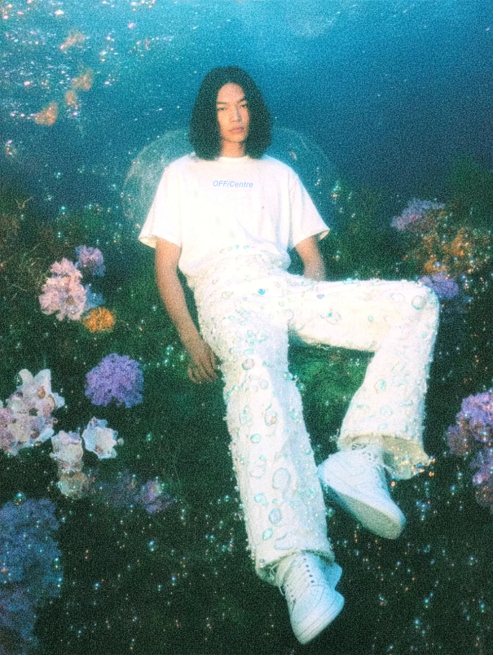 A person seated among vibrant flowers, wearing a white OFF/Centre t-shirt and embellished white pants. The background is a dreamy underwater scene with blue hues and sparkling elements.