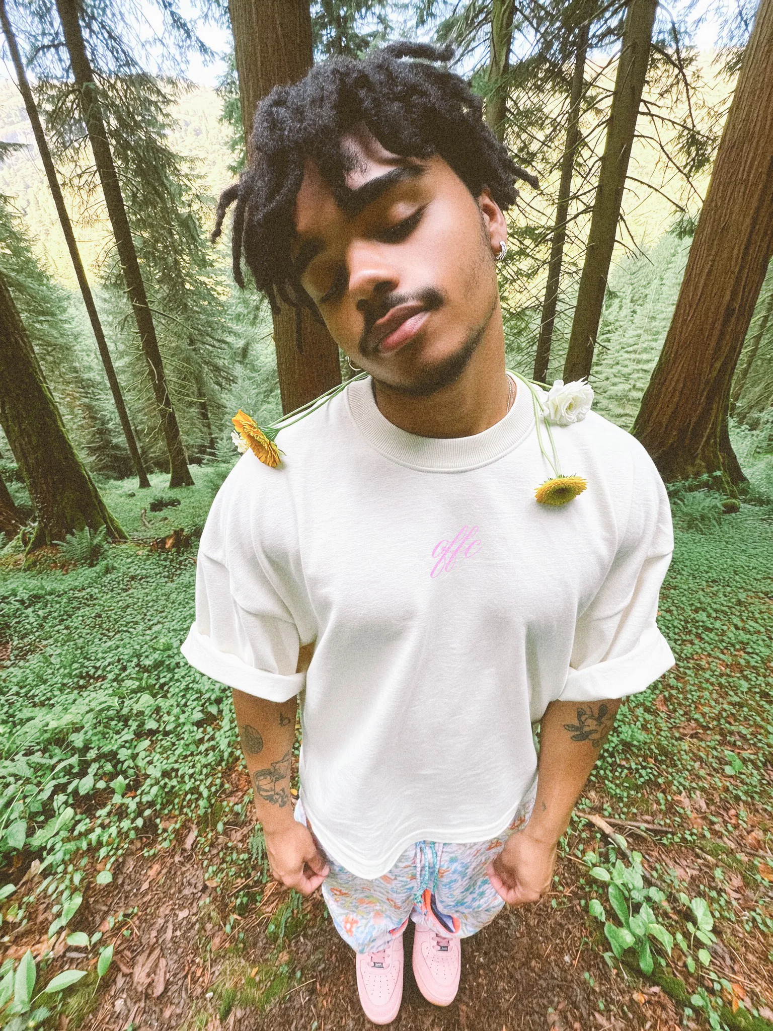 A person with dreadlocks standing in a forest, wearing a white shirt with floral decorations on the shoulders and patterned pants, with pink shoes.